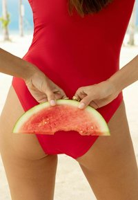 Rdeč enodelni kopalke iz raztegljive tkanine, z gladko teksturo. Roka drži klin vode melon, ki prikazuje njegovo zeleno lupino in sočno rožnato meso.