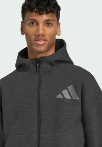 Sweat à zip gris avec capuche, fabriqué en tissu texturé. Il dispose d'un logo réfléchissant sur la zone de la poitrine gauche et de poches avant.