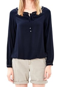 Blusa a maniche lunghe blu navy con collo rotondo, dettaglio con fiocco e tessuto testurizzato, abbinata a pantaloni corti beige chiaro arrotolati.