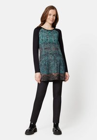 Langärmelige Tunika mit einem türkis-schwarzen Blumenmuster, schwarzen Ärmeln und entspannter Passform. Kombiniert mit schwarzen engen Hosen und klobigen schwarzen Stiefeln.