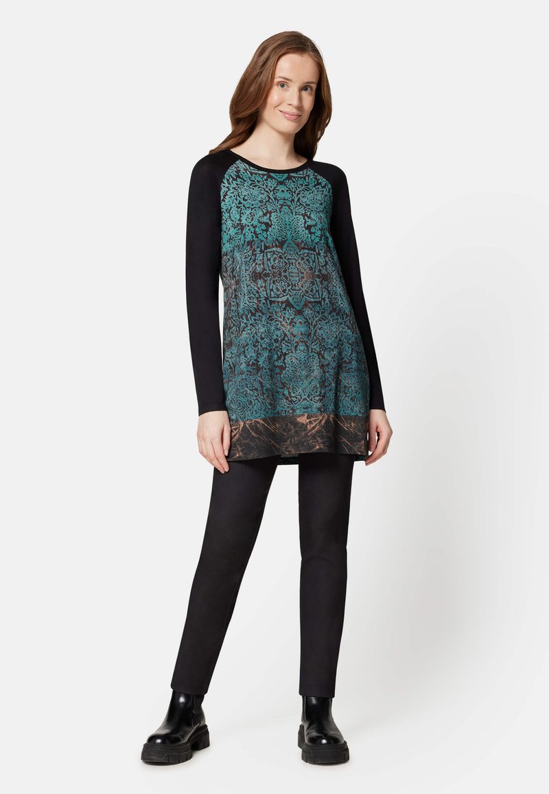 Langärmelige Tunika mit einem türkis-schwarzen Blumenmuster, schwarzen Ärmeln und entspannter Passform. Kombiniert mit schwarzen engen Hosen und klobigen schwarzen Stiefeln.