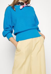 Femme portant un pull texturé bleu vif, un pantalon beige à jambes larges et une écharpe rouge, blanche et bleue nouée autour du cou.