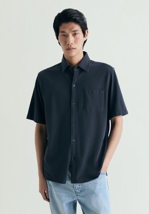 Joven con camisa negra de manga corta abotonada y vaqueros azul claro, de pie contra un fondo claro y liso.