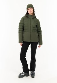 Groene pufferjack met een ritssluiting aan de voorkant, hoge kraag en een gepaneeld ontwerp. Gecombineerd met zwarte broek en zwarte skischoenen. Getextureerde gebreide muts.