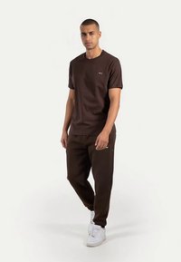 Brun bomulds t-shirt med korte ærmer, kombineret med matchende brune joggebukser med sidelommer, suppleret med hvide sneakers.