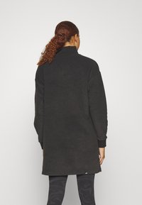Zwarte oversized fleece jas met een hoge kraag, lange mouwen en geribbelde manchetten. De stof heeft een gestructureerde afwerking. Fijne branding aan de zoom.