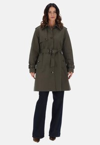 Trench coat verde oliva con vita cinturata, bottoni doppi, spalline e grandi tasche anteriori. Pantaloni in denim scuro a sbuffo.