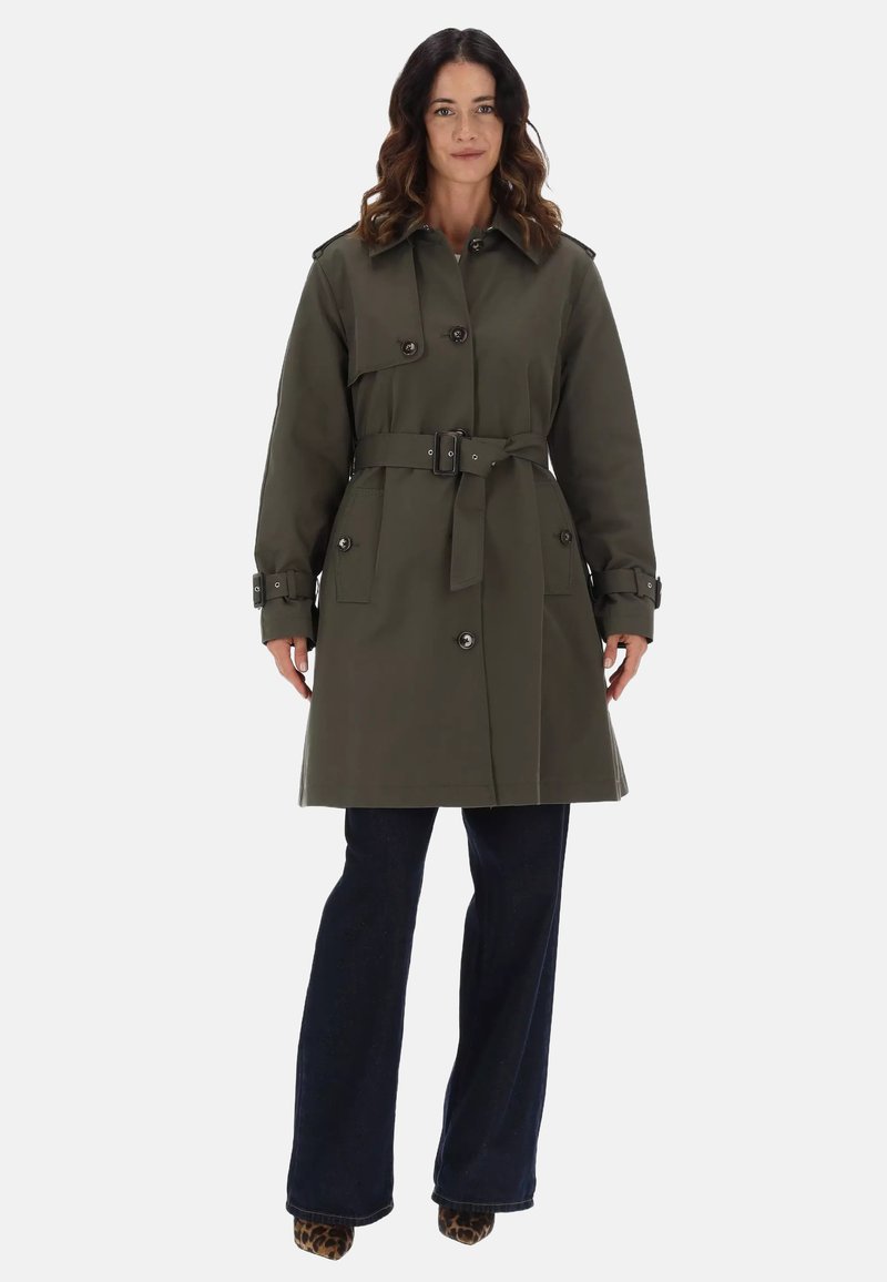 Trench coat verde oliva con vita cinturata, bottoni doppi, spalline e grandi tasche anteriori. Pantaloni in denim scuro a sbuffo.