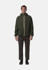 Giacca a felpa verde con cappuccio e zip frontale, due tasche anteriori e striscia multicolore lungo la zip; indossata con pantaloni e scarpe scuri.