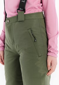 Groene broek van gladde stof, met een ritssluitingszak, lussen voor een riem en bijpassende zwarte bandjes. Gekocht met een roze top met lange mouwen.
