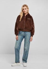Veste en fausse fourrure marron à zip avec capuche, poignets et ourlet côtelés, arborant un petit logo brodé. Associée à un jean bleu clair et des baskets blanches.