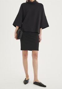 Černý oversized svetr s spadlými rukávy, kombinovaný s upnutou černou sukní. Modelka drží černou kabelku a má na sobě černé špičaté baleríny.