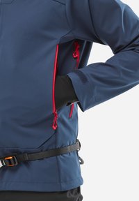 Personne portant une veste bleue avec une fermeture éclair rouge et des gants noirs, main dans la poche latérale, ceinture noire avec boucle orange autour de la taille.