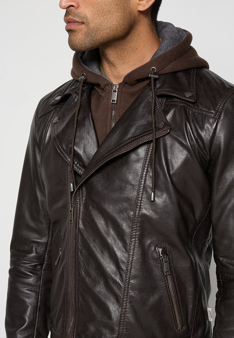 Chaqueta de motocicleta de cuero marrón con cremalleras plateadas, frente asimétrico y capucha con cordón. Presenta bolsillos con cremallera y acentos cosidos.