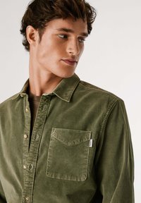 Camisa de pana verde oliva con cuello tipo botón, bolsillo frontal y costuras en contraste. Textura suave con un ligero brillo.