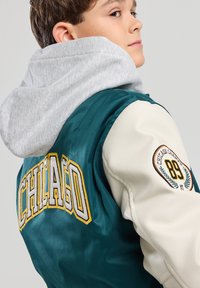 Green satin bomber jacket with cream sleeves, featuring "CHICAGO" in embroidered yellow and gray, and a gray hoodie underneath.