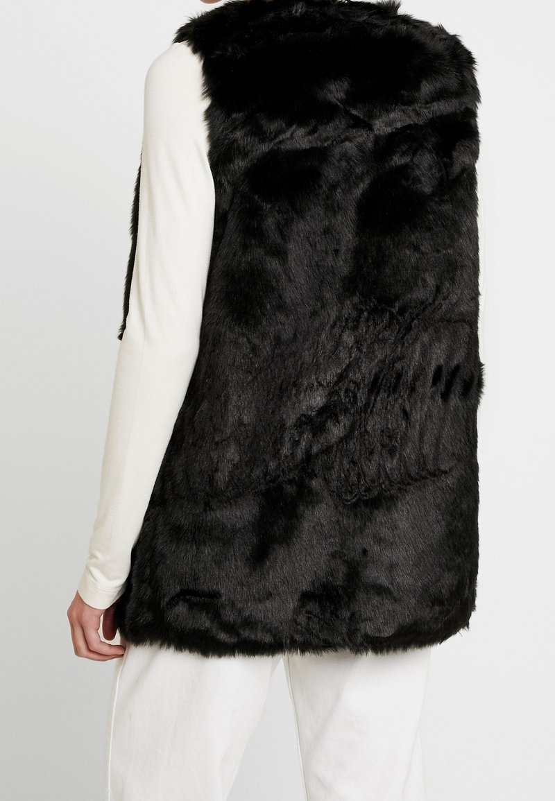 Svart fuskpälsväst med en mjuk textur, med ärmlös design och rak skärning, bärs över en ljusfärgad topp.