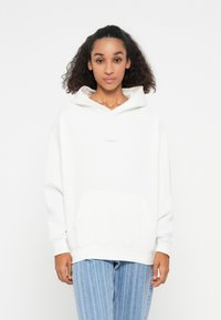 Weißer Hoodie aus weichem Stoff mit einer Kängurutasche, gerippten Bündchen und einem kleinen Logo auf der Brust. Kombiniert mit hellblauen gestreiften Jeans.