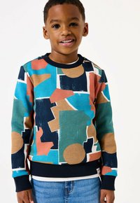 Ragazzino sorridente che indossa un maglione con motivo geometrico colorato in blu, teal, beige e corallo, abbinato a jeans blu, su uno sfondo bianco.