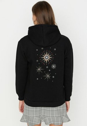 Femme portant un sweat à capuche noir orné de motifs de soleil et d'étoiles sur le dos, associée à une jupe plissée grise à carreaux, vue de dos.