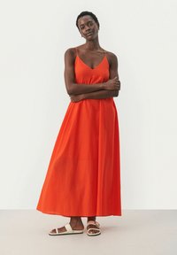 Lång, ärmlös, klarorange klänning med åtsittande liv och svävande kjol. Bärs med vita platta sandaler med en texturerad tyg.