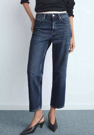 Vrouw draagt donkerblauwe jeans met rechte pijpen, zwarte slingback hakken en een zwart topje met lange mouwen, staand tegen een witte muur.