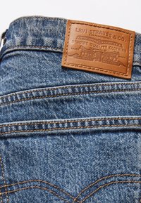 Mörkblå denim jeans med en brun läderetikett som har präglade detaljer vid midjan. Kraftig sömnad synlig längs sömmarna.