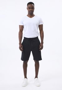Camiseta blanca de cuello en V, pantalones cortos negros texturizados con cinturilla elástica y zapatillas blancas. El modelo está de frente con un fondo neutro.