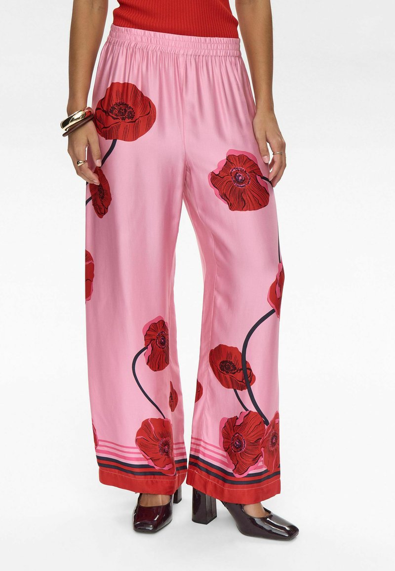 Pantaloni larghi in satin rosa con motivo floreale a papaveri rossi, vita elasticizzata e orlo a righe in contrasto. Abbinati a scarpe con tacco scuro.