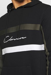 Sudadera negra con capucha que presenta un bolsillo frontal, franjas en contraste verde oliva y blanco, con la palabra "Closure" en letras blancas cursivas en el pecho.