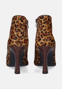Botas de tobillo con estampado de leopardo y acabado de piel texturizada, detalle de cremallera negra y tacón de aguja de madera. Patrón audaz sobre fondo marrón claro.