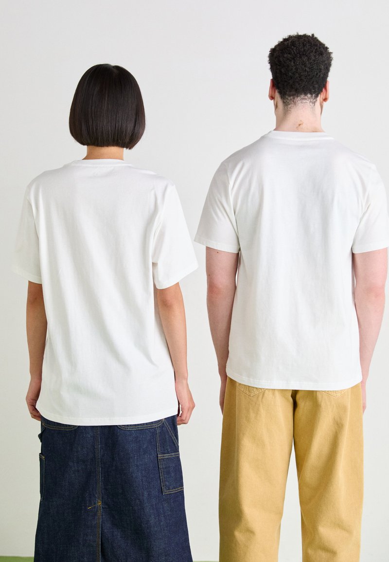 Dos personas vestidas con camisetas blancas lisas, de pie espalda con espalda. Una lleva vaqueros oscuros y la otra pantalones amarillos claros.