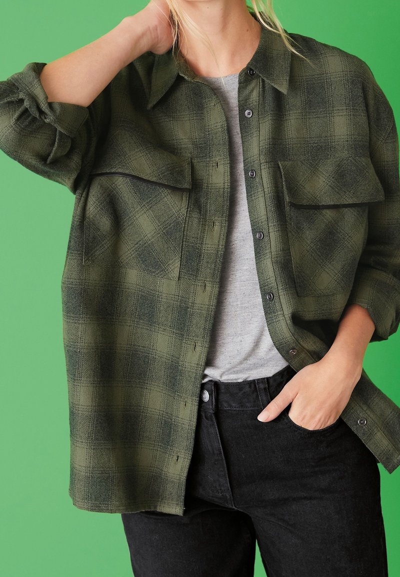Personne portant une chemise à carreaux verte boutonnée sur un t-shirt gris, avec une main dans la poche d'un jean noir et l'autre touchant son cou.