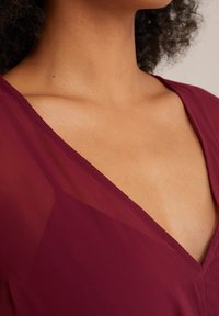 Burgundy top featuring a deep V-neckline with sheer fabric overlay and soft, smooth texture. The cut accentuates the collarbone.