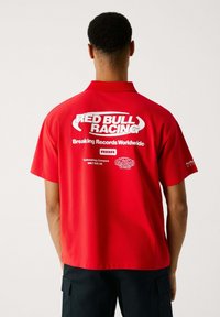 Sarkans polo krekls ar īsām piedurknēm, uz muguras ar uzrakstu "RED BULL RACING" un "Breaking Records Worldwide" baltā krāsā. Gluda auduma.