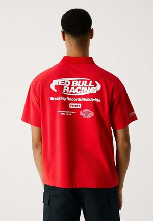Röd polo med korta ärmar, med "RED BULL RACING" och "Breaking Records Worldwide" i vitt på baksidan. Slät tyg.