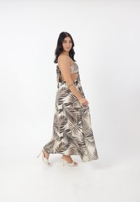 Maxikleid mit einem braunen und cremigen Palmenblattmuster, trägerlosem Design, offenem Rücken und strukturiertem Stoff. Kombiniert mit sandaletten mit Absatz.