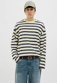 Camisa de manga larga a rayas en color crema y marino, hecha de algodón, combinada con jeans de color azul claro y un cinturón marrón. Una gorra casual realza el atuendo.