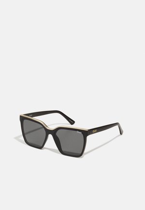 Black sunglasses with a thick frame, gold accents along the top edge, and dark lenses. Features round corners and "QUAY" branding on the arms.