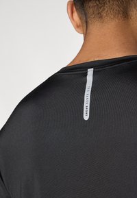 Čierne športové tričko s hladkou textúrou, na zadnej strane s bielou vertikálnou etiketou s nápisom "TED BAKER SPORT".