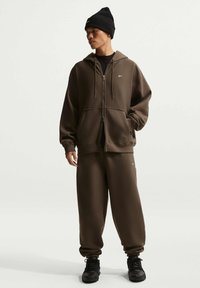 Hombre con chándal Nike marrón oscuro que incluye sudadera con capucha con cremallera y pantalones deportivos, gorro negro y zapatillas negras, de pie contra un fondo liso.