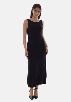 Donna che indossa un vestito nero senza maniche, lungo fino al pavimento, con tacchi neri appuntiti, in piedi davanti a uno sfondo chiaro e uniforme.