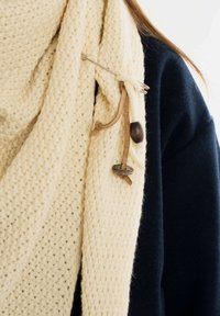 Kremowy, dziany szalik o teksturowanym wzorze, z drewnianym guzikiem i skórzanymi akcentami, nałożony na ciemnoniebieską odzież.