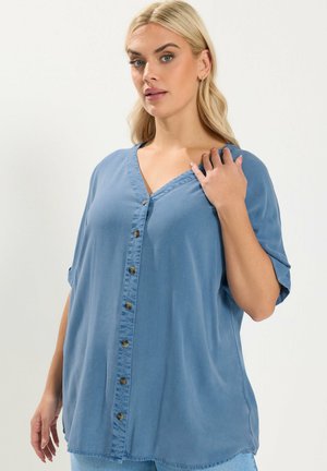 Donna bionda che indossa una camicia blu larga con bottoni e maniche arrotolate, che posa con una mano vicino al colletto su uno sfondo semplice.