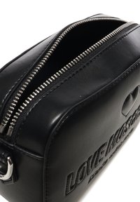 Borsetta cosmetica in pelle nera con testo in rilievo, zip argentata e una superficie liscia; forma rettangolare compatta con un design minimalista.