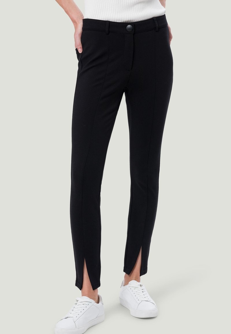 Femme portant un pantalon noir slim avec des fentes à l'avant aux chevilles, mains dans les poches, baskets blanches et un haut blanc côtelé.