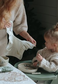 Silikonebibs med hvide, flæsekantede kanter og udskårede mønstre, holdt af en voksen. Barn i en striktrøje sidder ved et bord med service tæt ved.