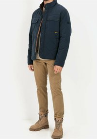 Marineblauwe gewatteerde jas met twee borstzakken en bruine voering, gecombineerd met beige broeken en tan laarzen. Heeft een klassiek kraagontwerp.