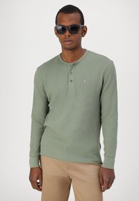 Zöld, hosszú ujjú henley póló ribbed textúrával, kerek nyakkal és három gombos gallérral. Világosbarna nadrággal párosítva.