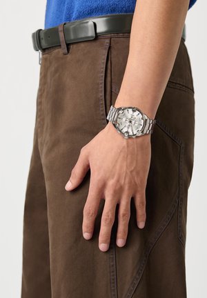 Pols met zilverkleurig metalen chronograafhorloge bij een persoon die een bruine broek en een zwarte riem met een blauw overhemd draagt.